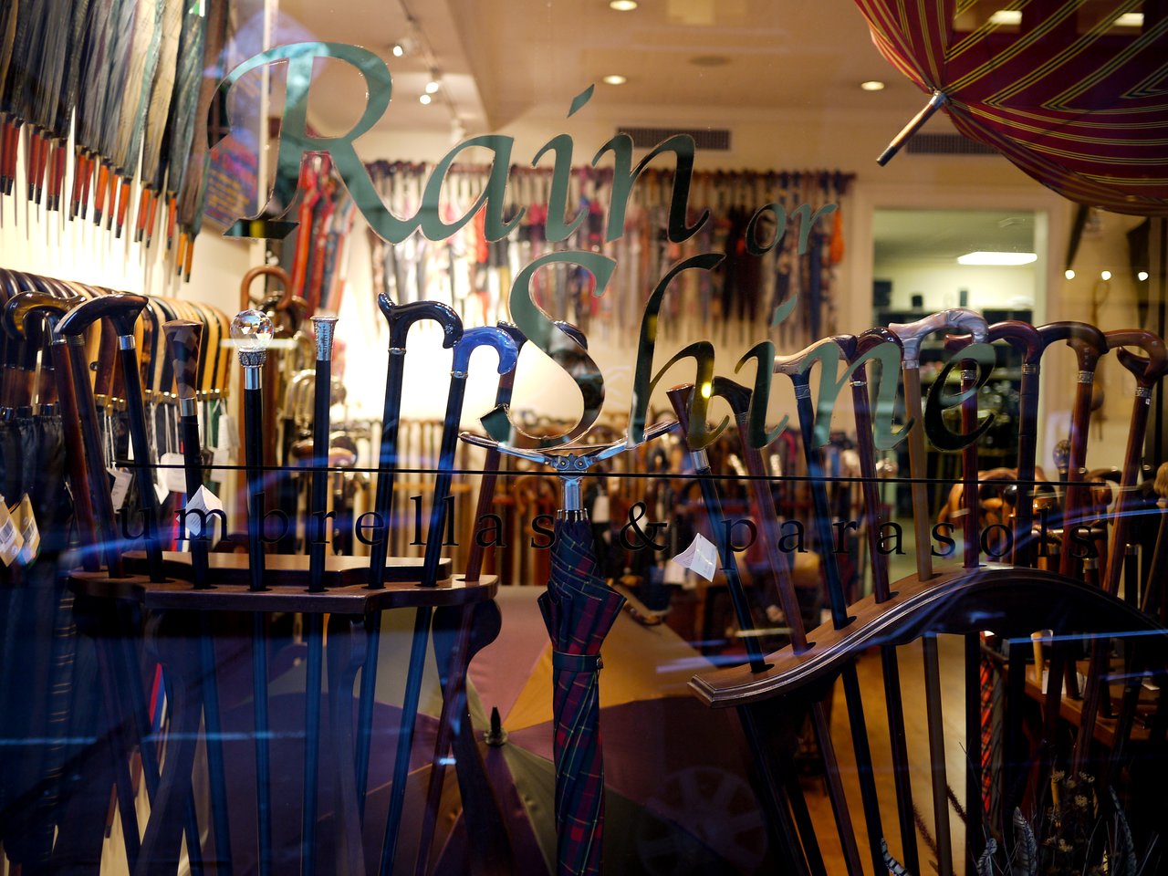 A store window displaying a variety of umbrellas and walking canes, with "Rain or Shine" written on the glass.