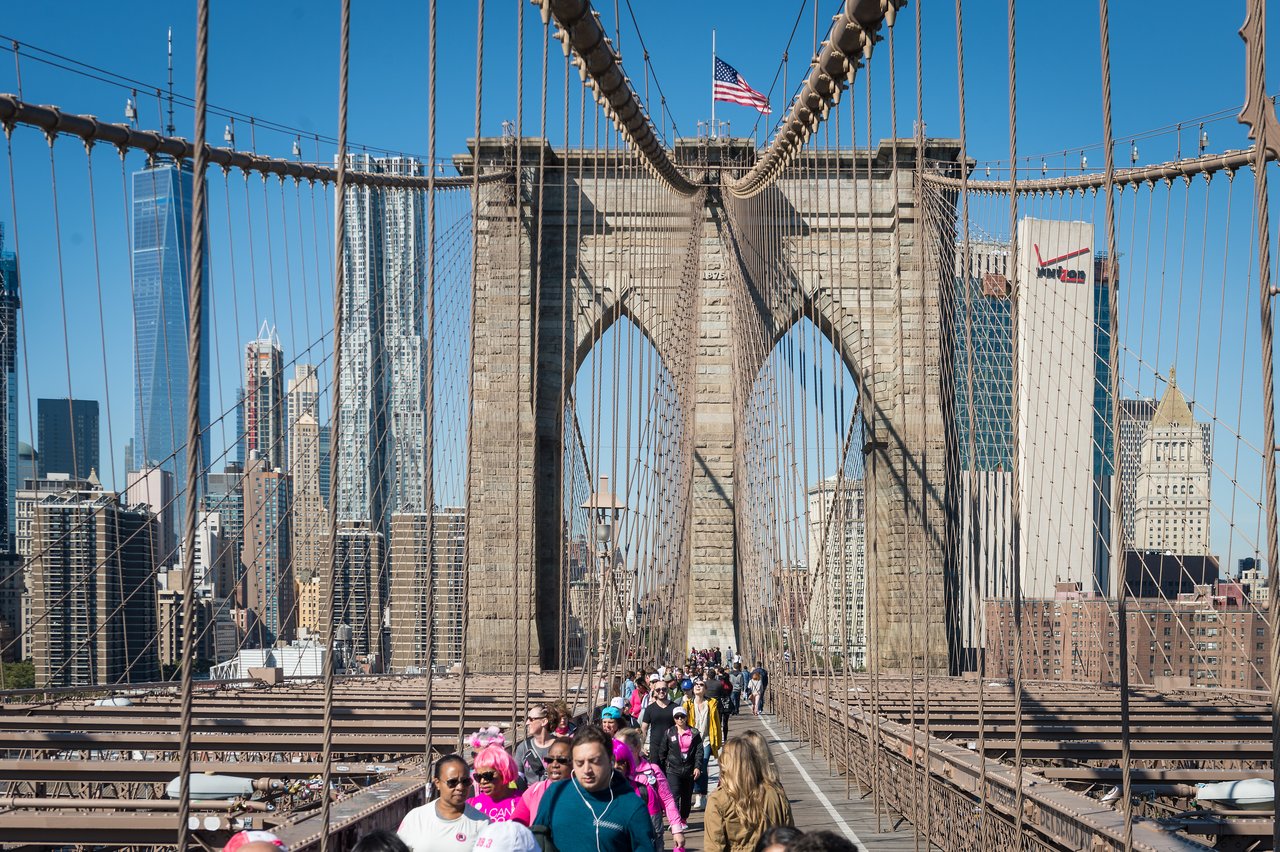 People walking on the Brooklyn Bridge with the New York City skyline in the background on a clear day.