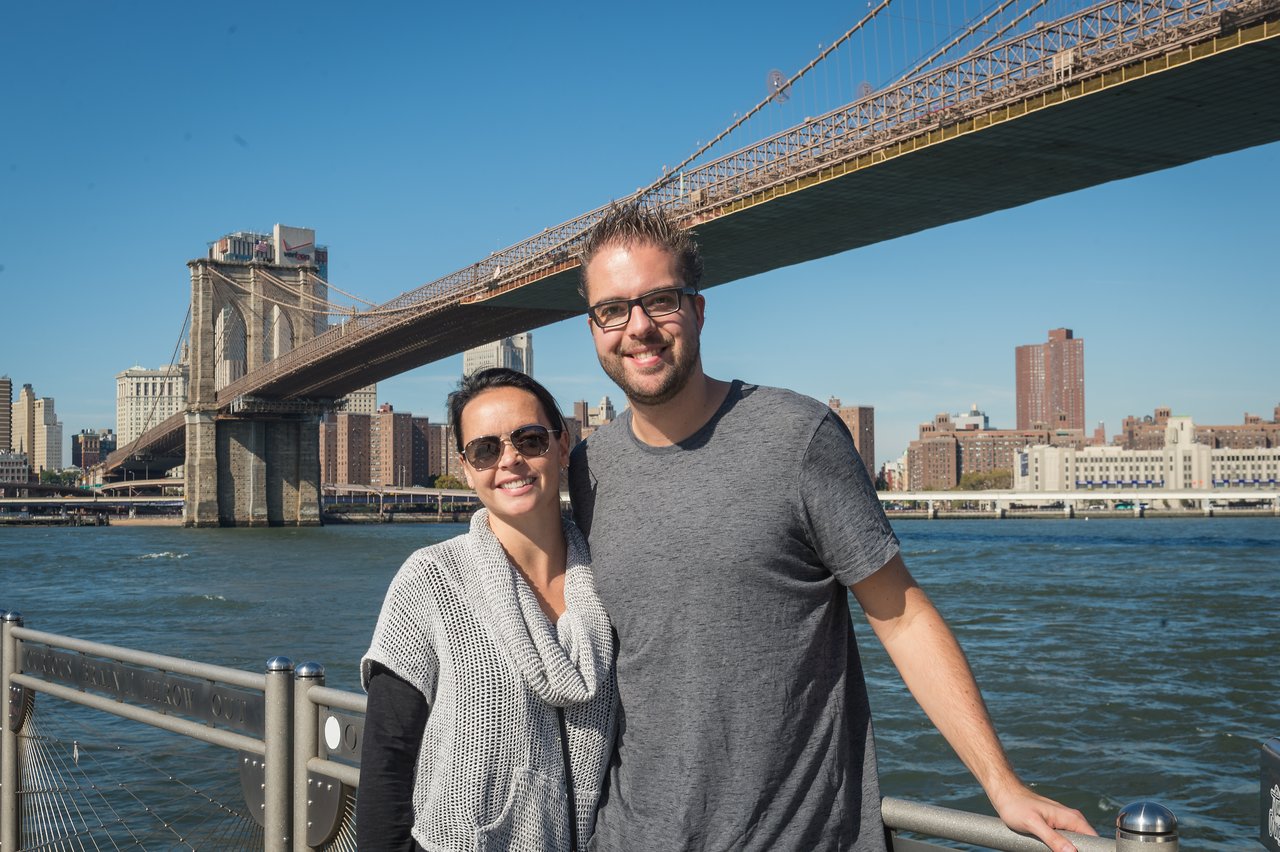 A smiling couple poses in front of the Brooklyn Bridge on a sunny day.