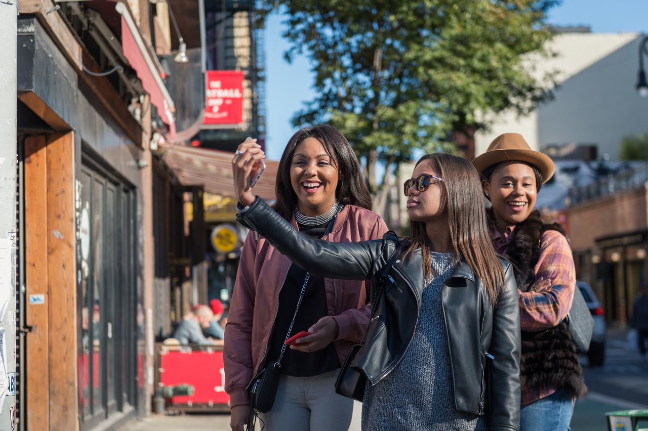 Three friends stand on a city street, smiling as one holds up a phone to take a selfie.
