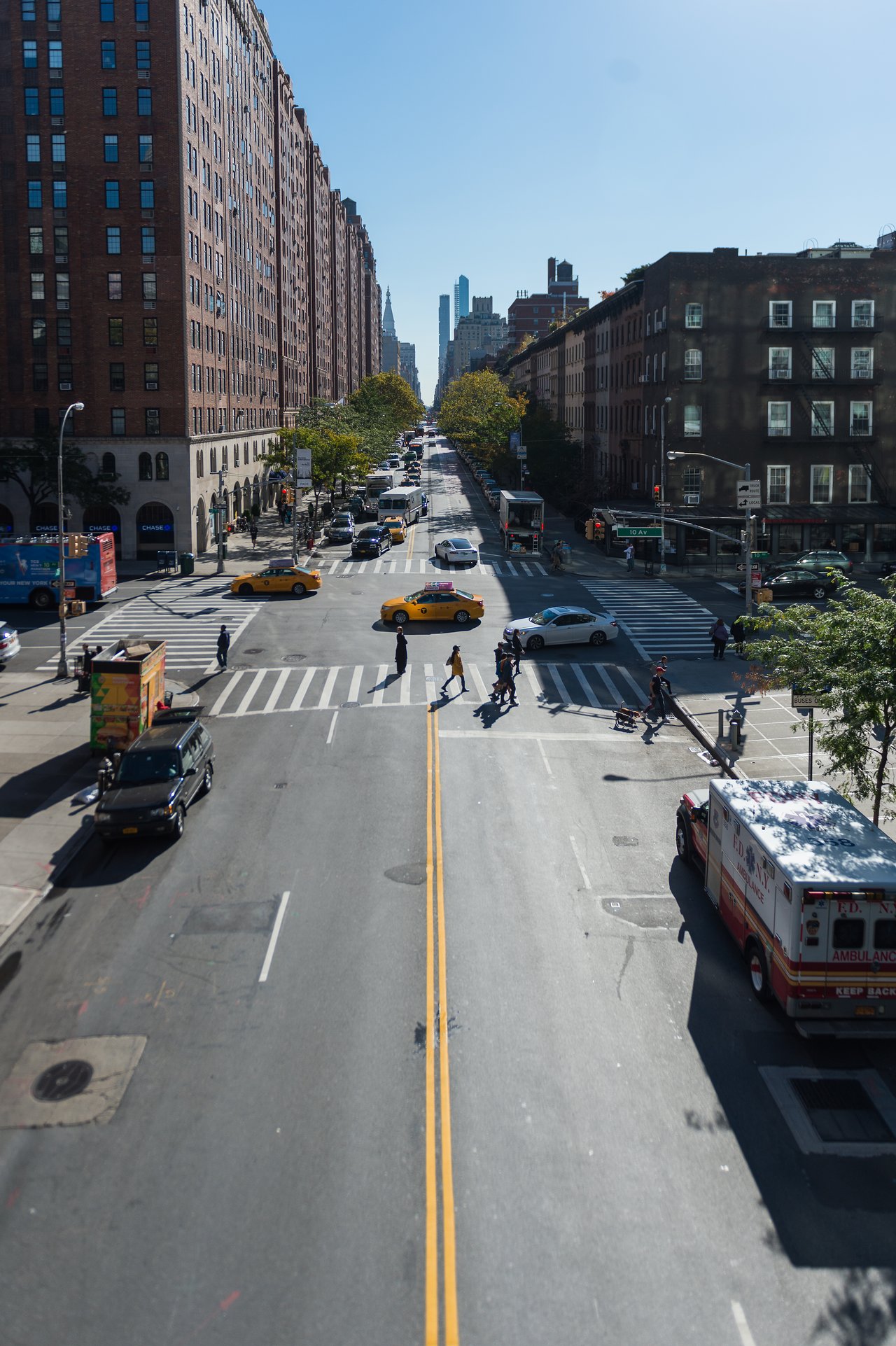 People cross a busy New York street with taxis, cars, and an ambulance stopped at the intersection.