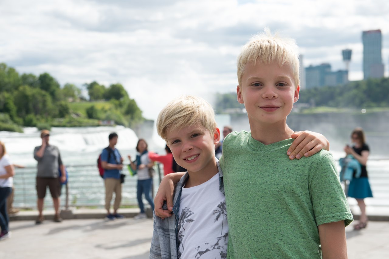 Two smiling boys stand close together, one with his arm around the other, posing for a photo outdoors.