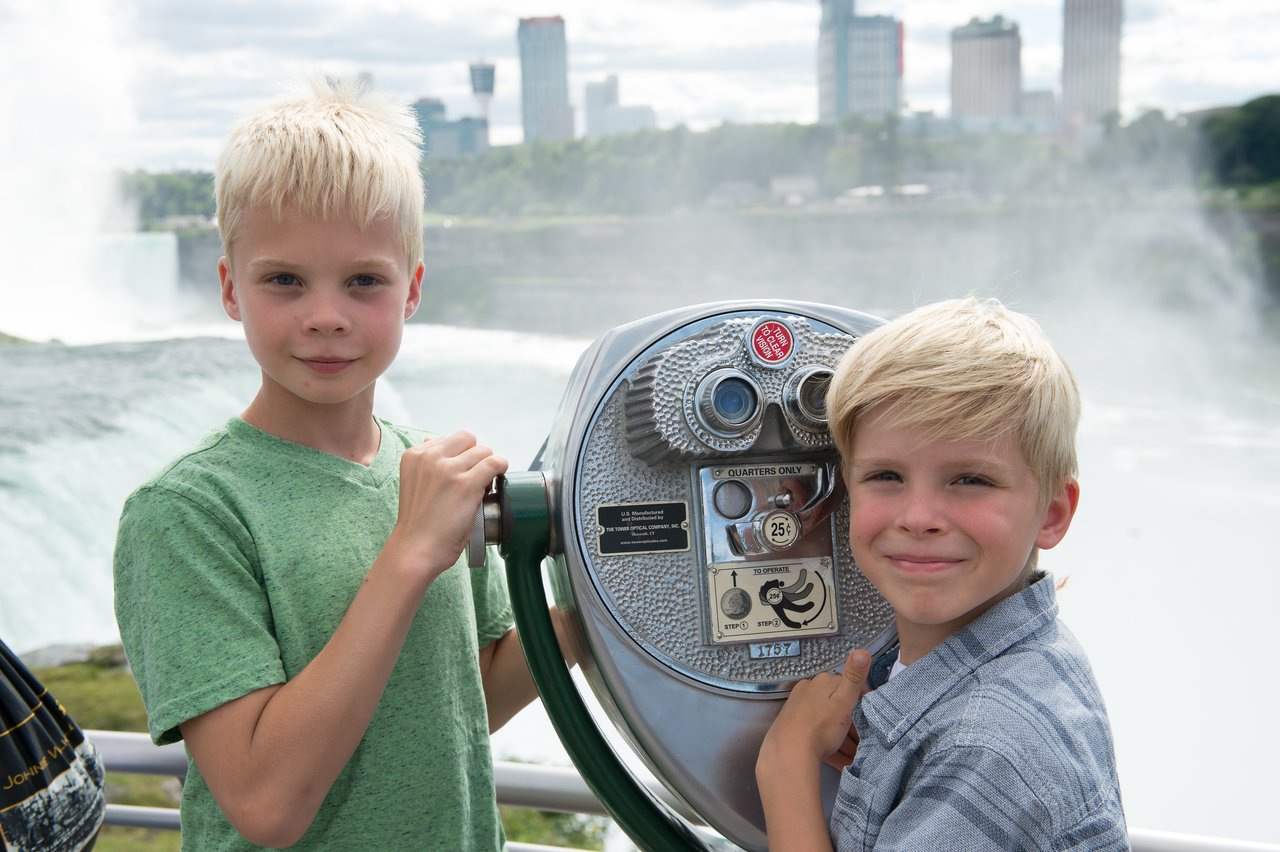 Two young boys stand next to a coin-operated viewing telescope, smiling at the camera.