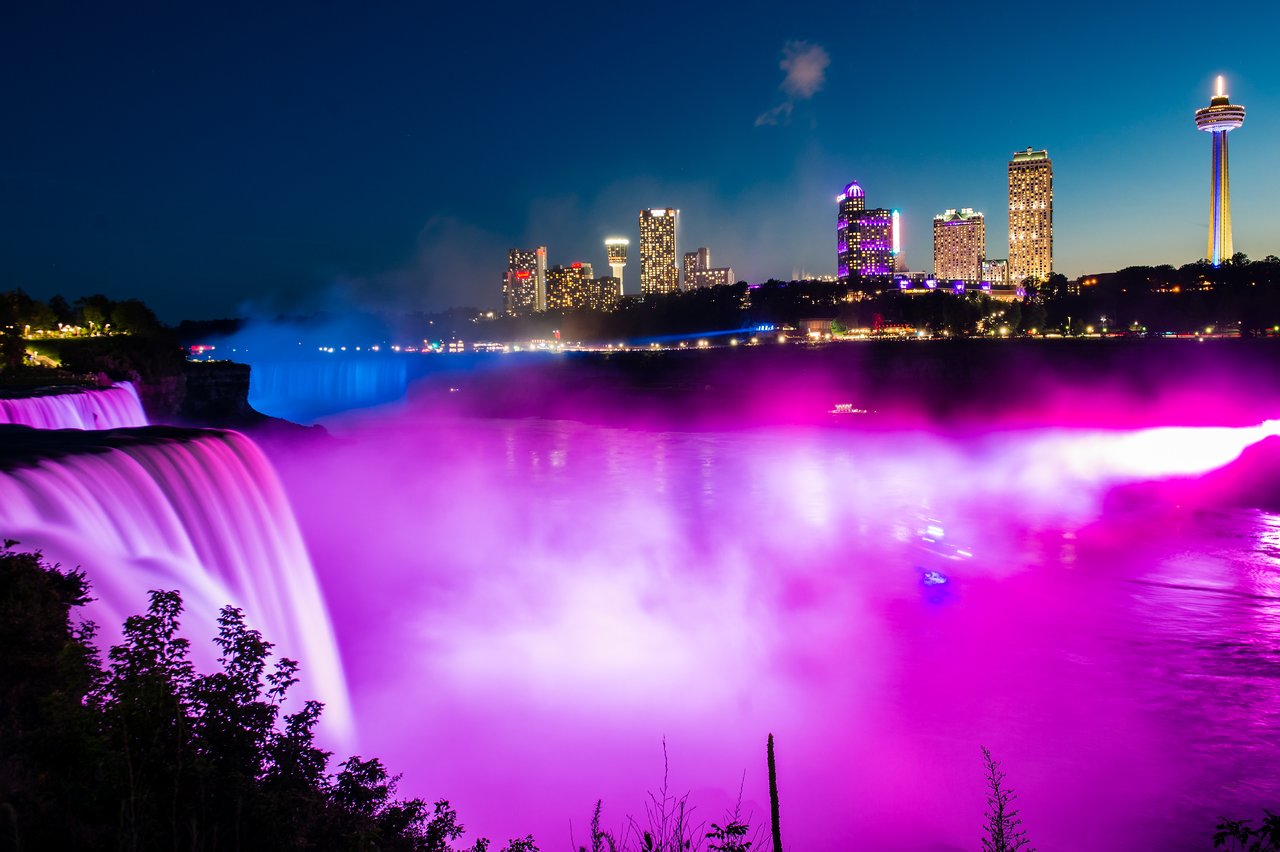 Niagara Falls glows with pink and purple lights at night, with a city skyline in the background.