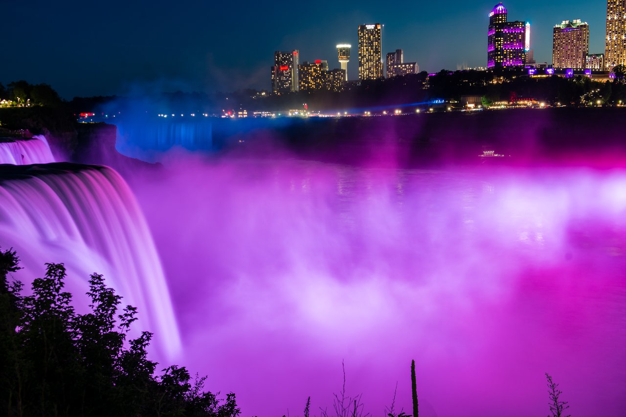 Niagara Falls at night, illuminated in purple and pink lights, with mist rising and a city skyline in the background.
