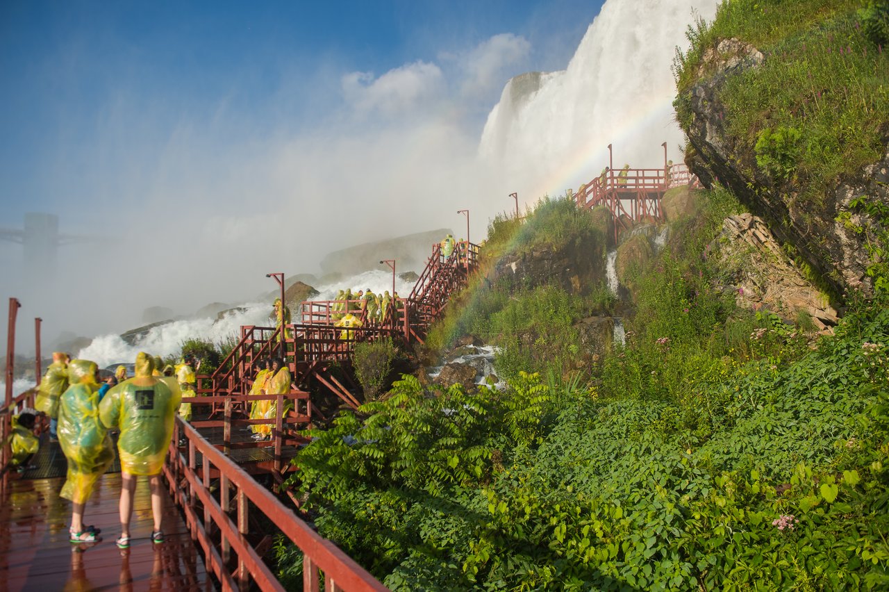 People in yellow rain ponchos walk on a wooden platform near a waterfall, with mist and a rainbow visible.