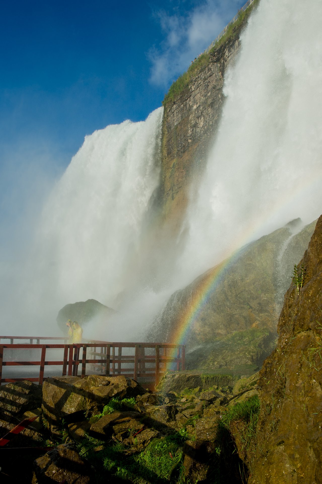 Two people in yellow raincoats stand on a wooden platform near a large waterfall, with a rainbow visible.