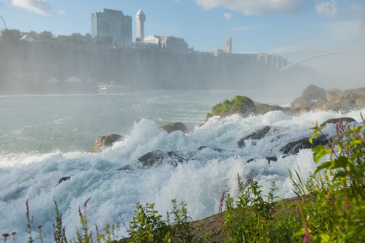 Rushing water flows over rocks at Niagara Falls, with mist rising and buildings visible in the background.