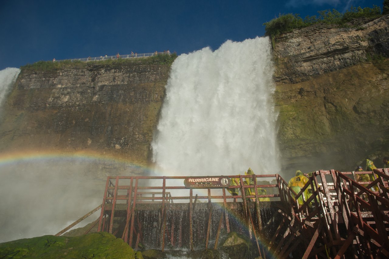 Visitors in yellow ponchos stand on a wooden platform near a powerful waterfall, with a rainbow in the mist.
