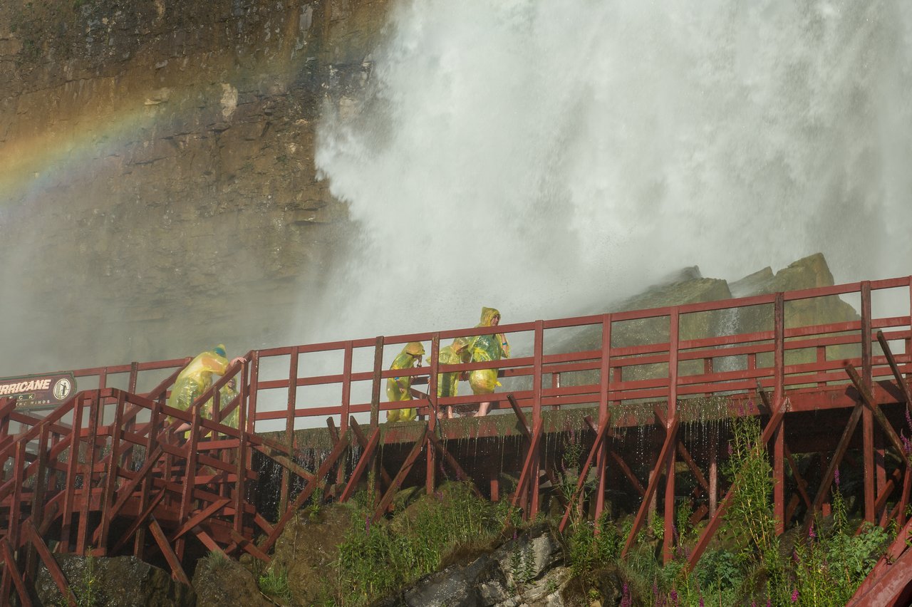 People in yellow rain ponchos walk on a wooden platform near a powerful waterfall, with water spraying around them.