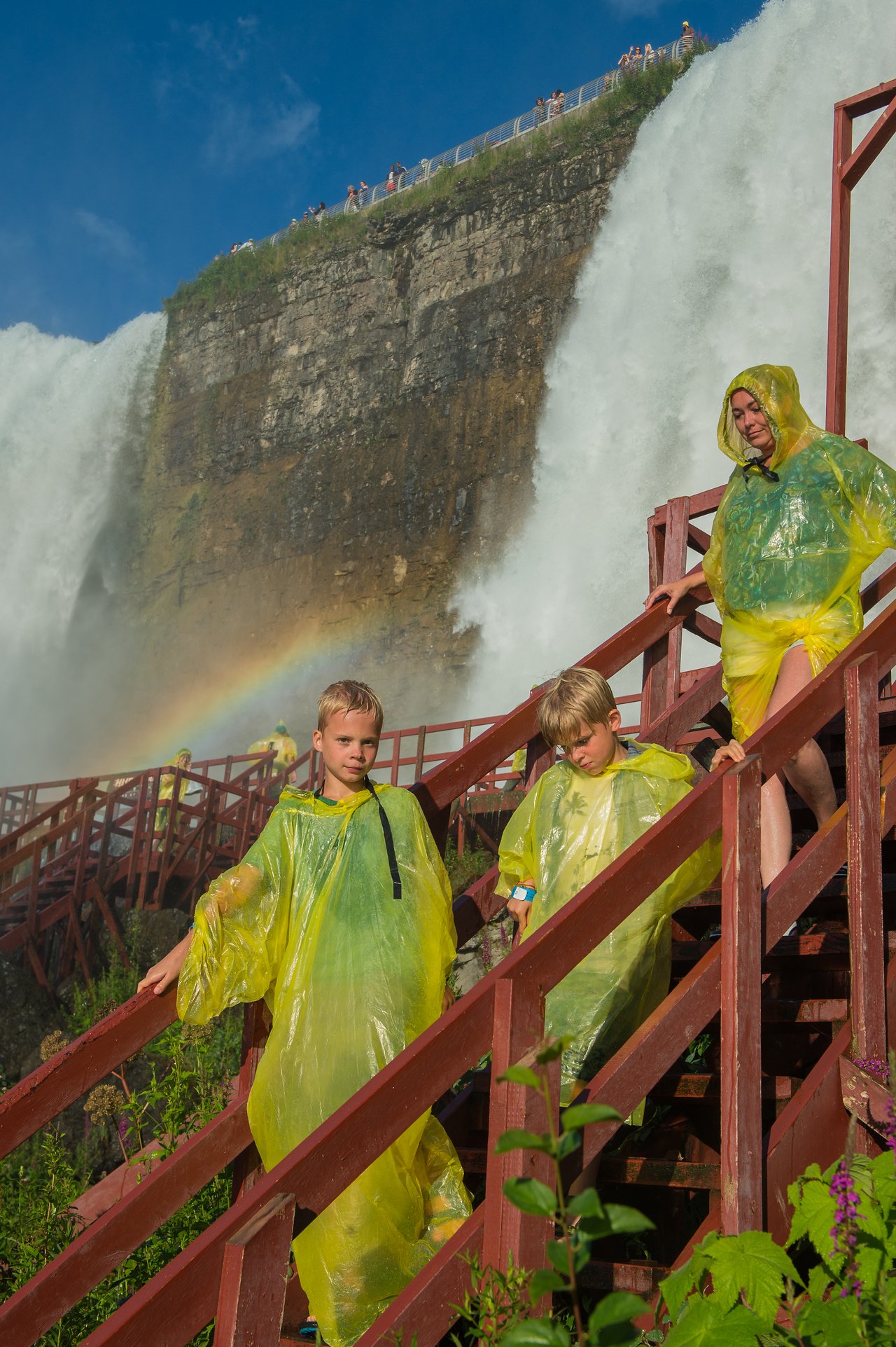 Three people in yellow rain ponchos walk down wooden stairs near a large waterfall.