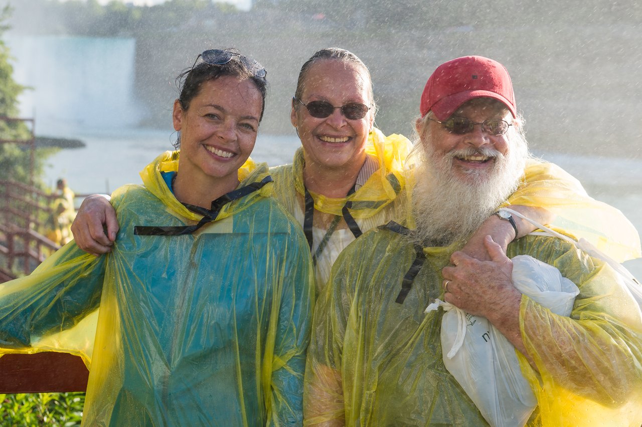 Three people wearing yellow rain ponchos smile while standing close together in a misty outdoor setting.