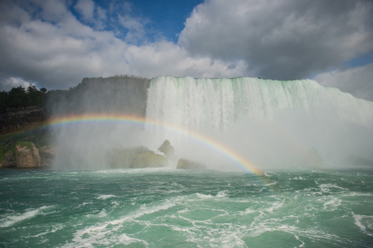 A large waterfall cascades down rocks with mist rising, creating a rainbow over the rushing water below.