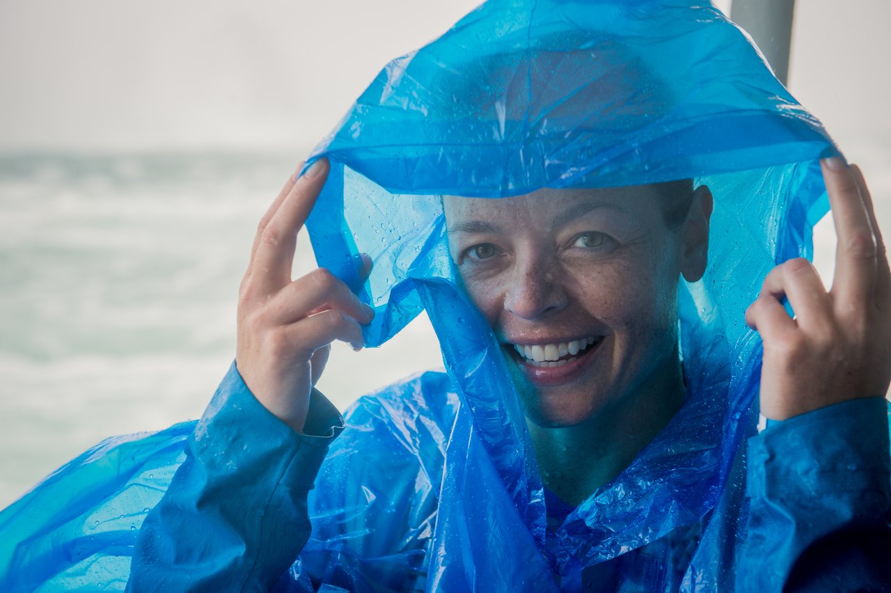 Smiling person wearing a blue rain poncho, holding up the hood with both hands.