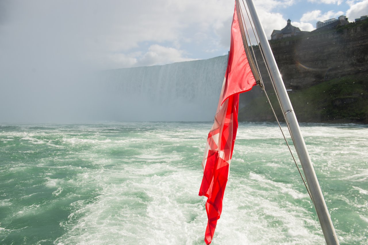 A Canadian flag flutters on a boat near a large waterfall with mist rising from the water.