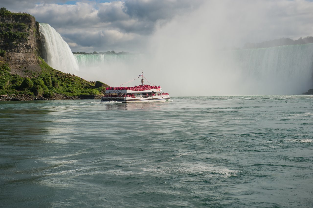 A tour boat filled with people in red raincoats approaches a large waterfall, surrounded by mist and water.