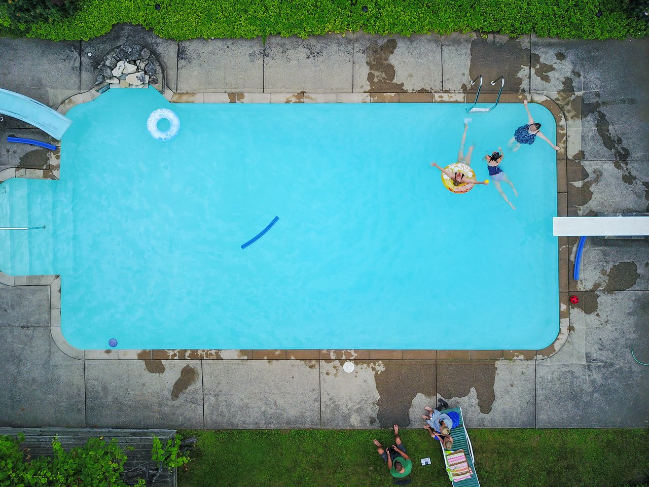Three people swim in a pool, one using a float, while two others sit on lounge chairs nearby.