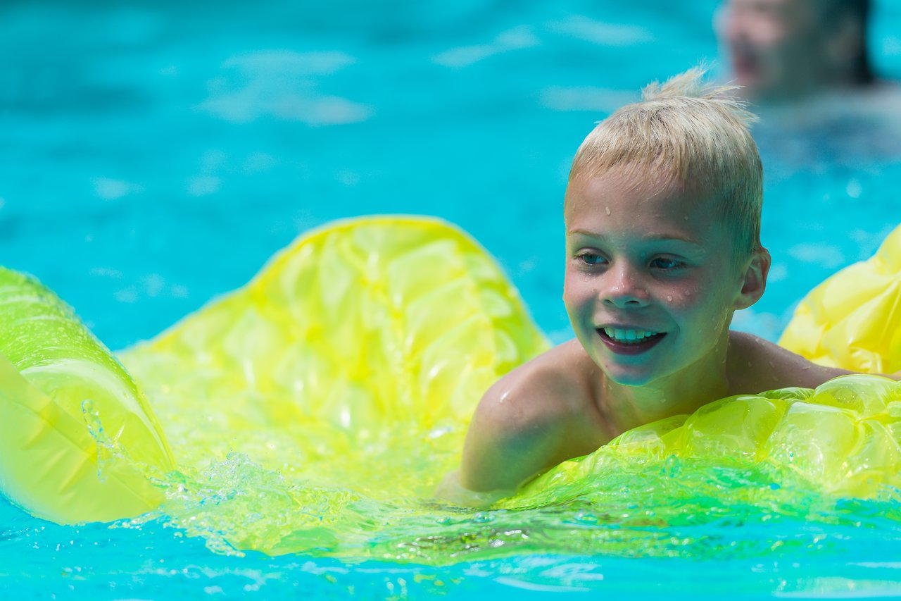 A smiling child floats on a yellow inflatable raft in a swimming pool, splashing water around.