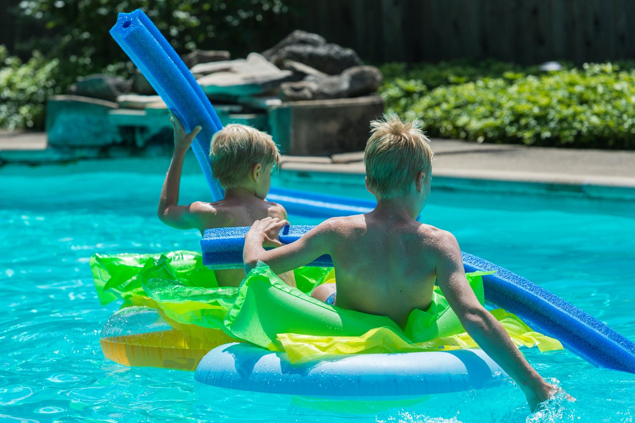 Two children sit on inflatable pool floats, holding foam noodles and playing in the water.