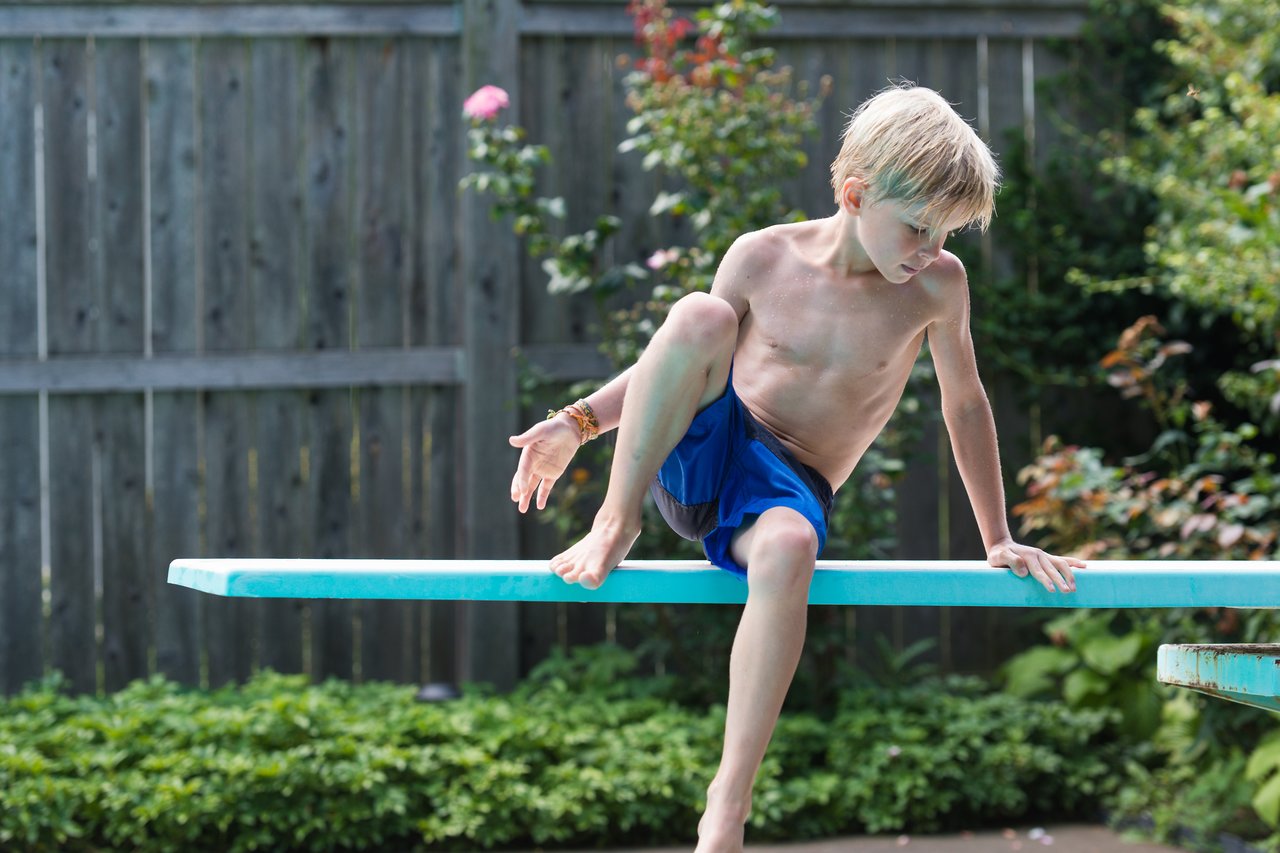 A boy in blue swim trunks climbs onto a diving board, preparing to jump into the pool.