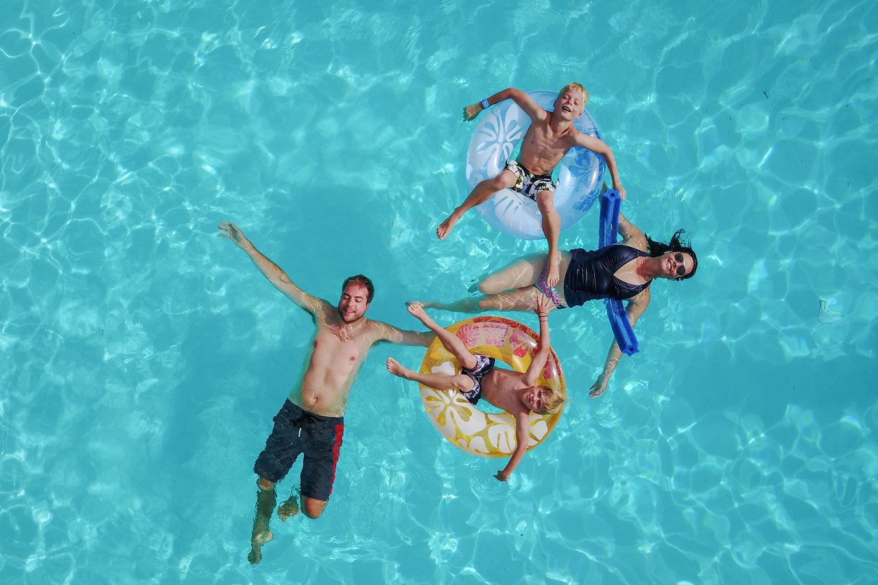 A family of four floats in a swimming pool, with two children in inflatable rings and parents relaxing nearby.