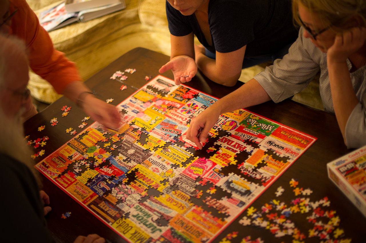 Four people work together to assemble a colorful jigsaw puzzle on a wooden table.
