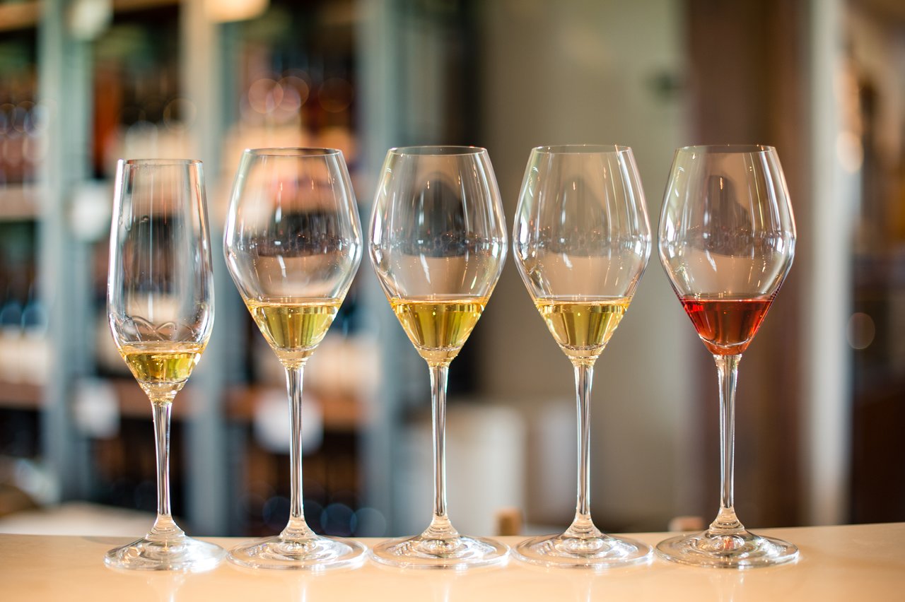 Five wine glasses with different amounts of white and red wine are lined up on a table.