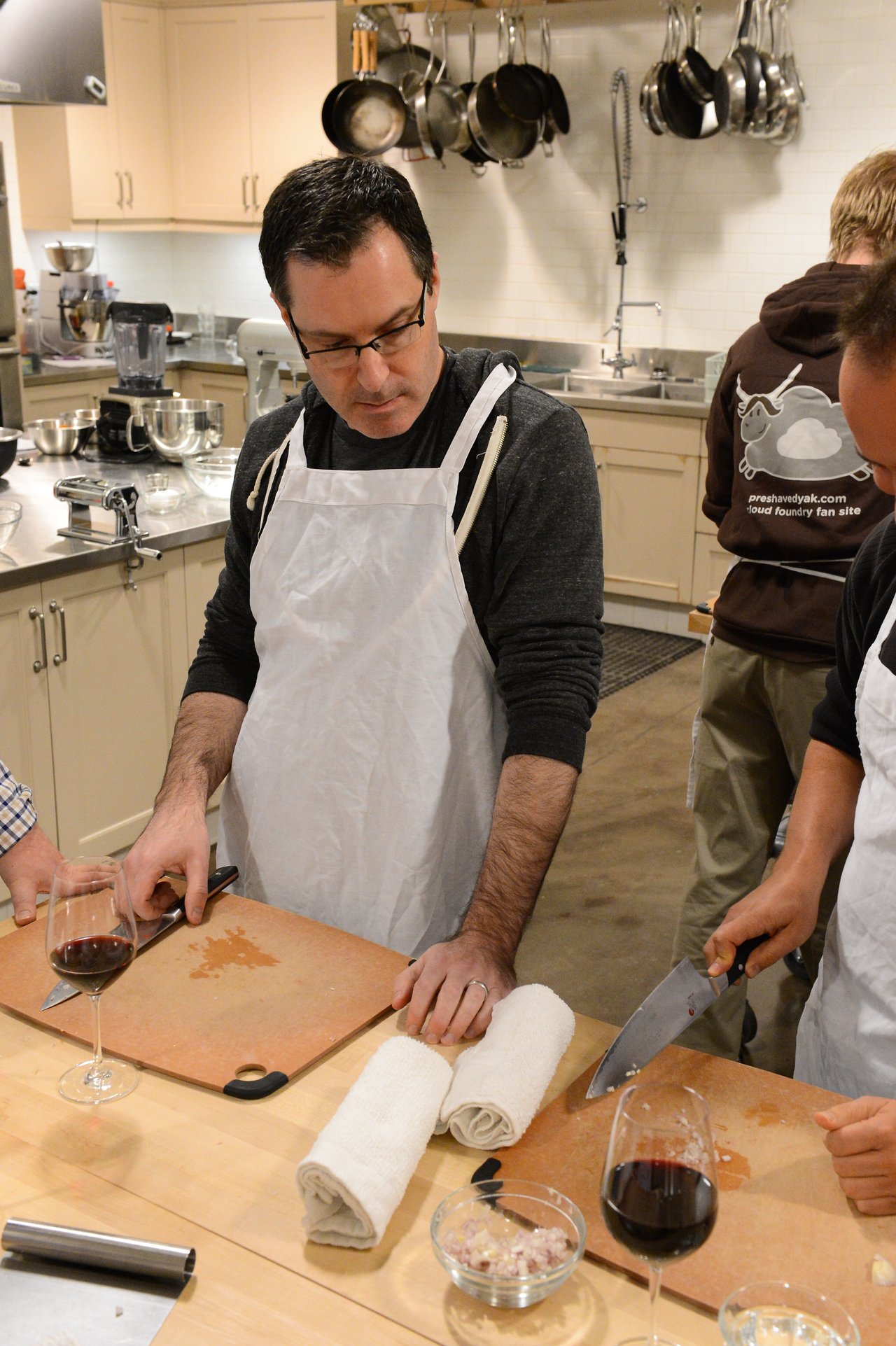 A man in an apron observes as another person chops ingredients on a cutting board in a cooking class.