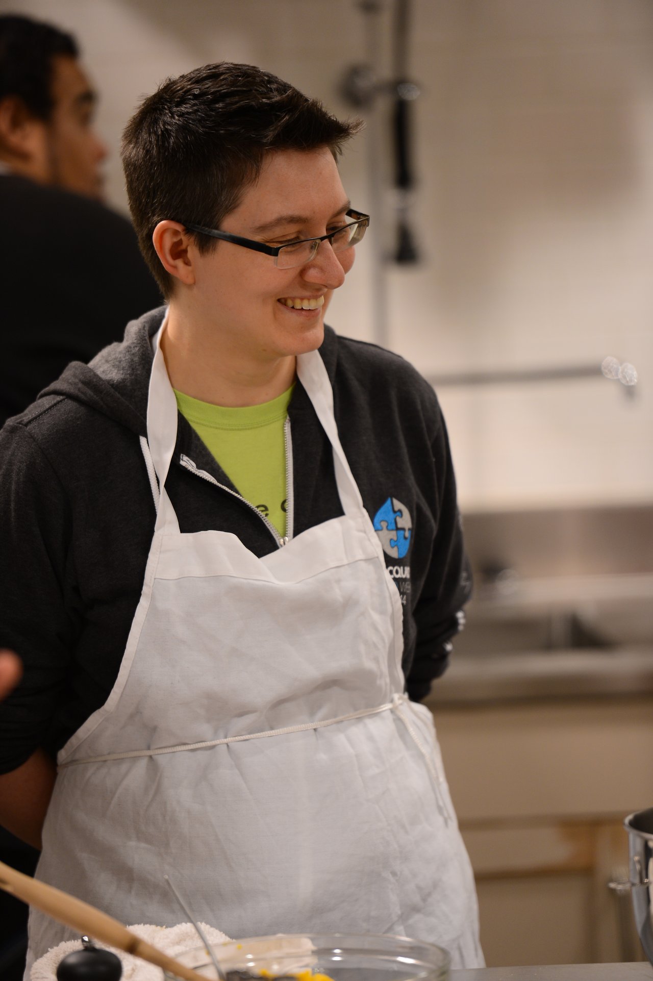 A person wearing glasses and a white apron smiles while participating in a cooking class.