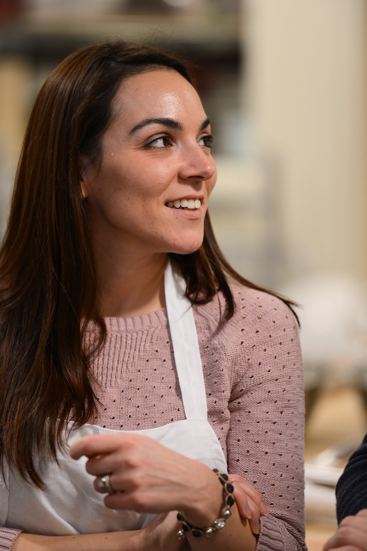 A woman wearing a white apron smiles and looks to the side during a cooking class.