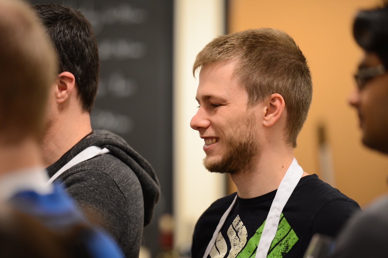 A group of people wearing aprons participate in a cooking class, with one person smiling and engaged in conversation.