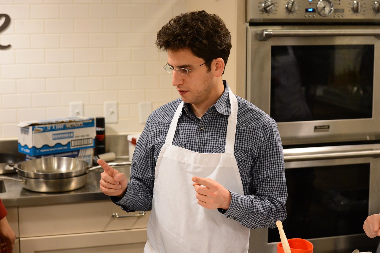A person wearing a white apron gestures while speaking in a kitchen during a cooking class.