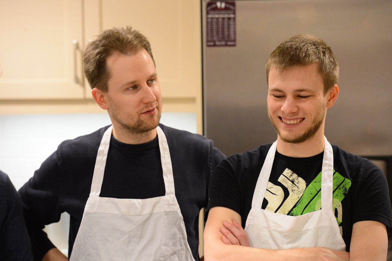 Two men wearing aprons stand in a kitchen, one smiling while the other speaks to him.