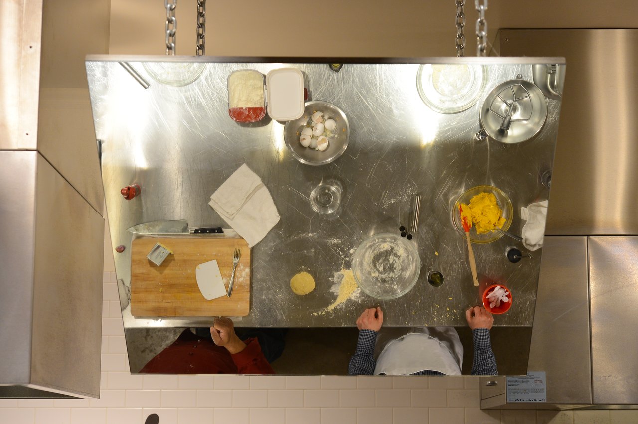 Two people preparing food at a stainless steel kitchen counter with ingredients, utensils, and a cutting board.