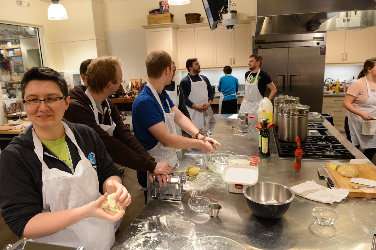 A group of people in aprons prepare dough and cook together in a kitchen during a cooking class.