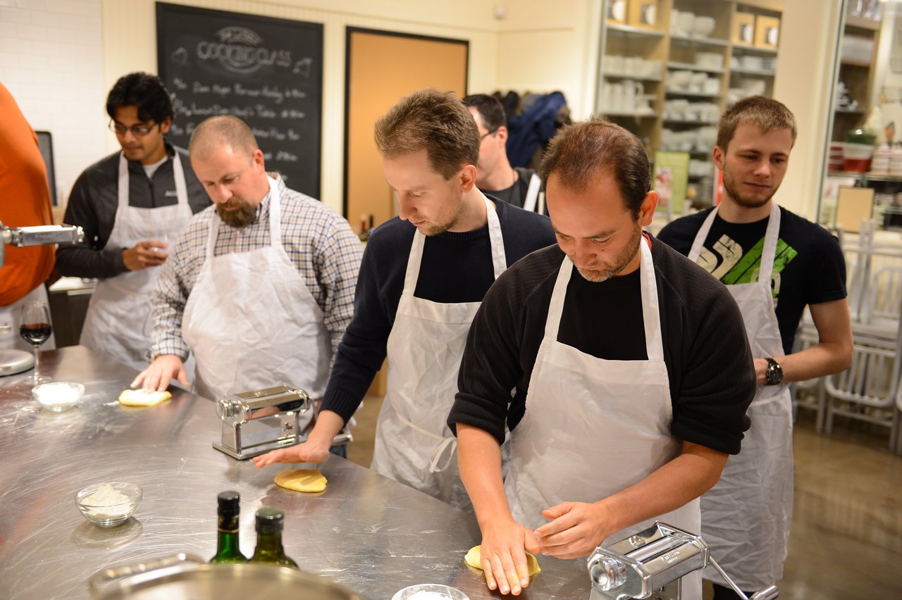 A group of people wearing aprons prepare dough at a cooking class, using pasta machines on a stainless steel table.