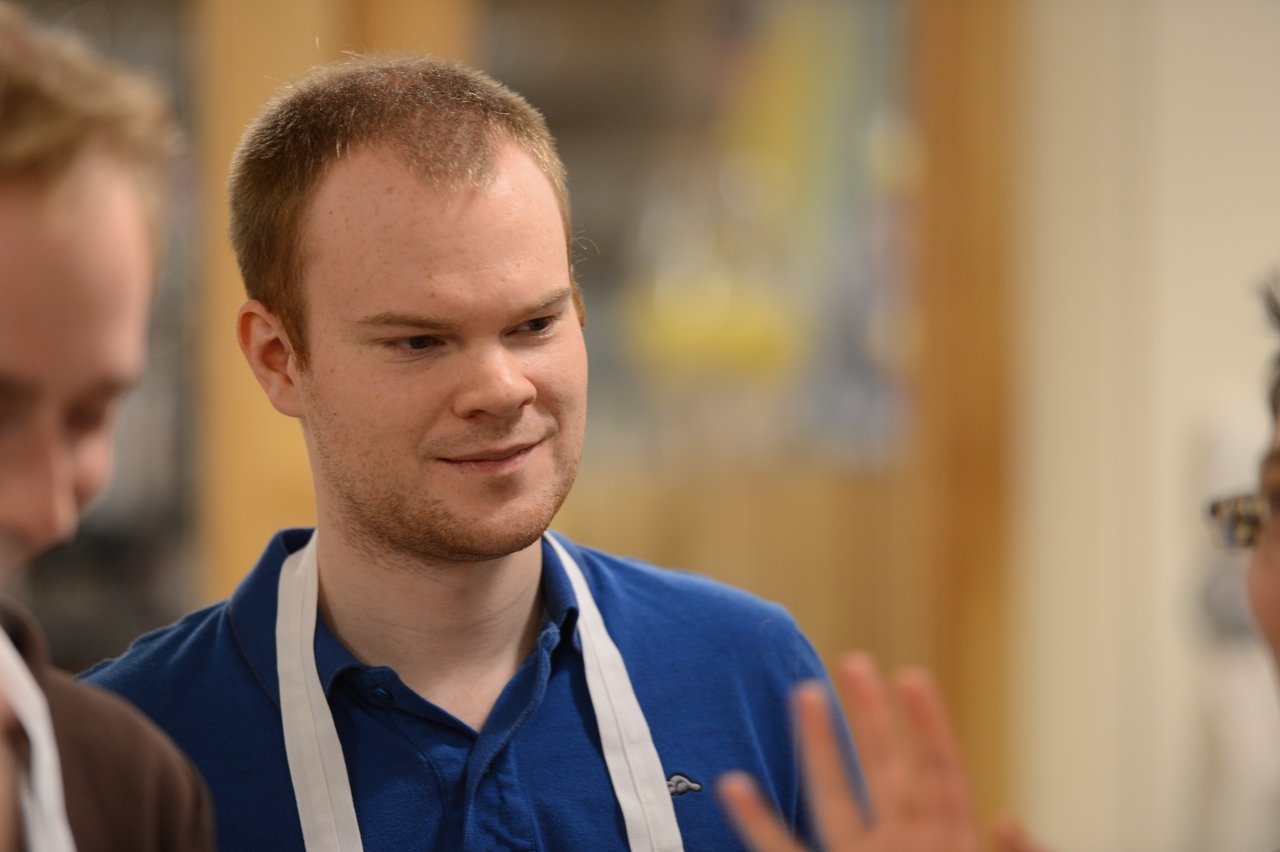 A man wearing a blue shirt and white apron listens while engaging in conversation during a cooking class.