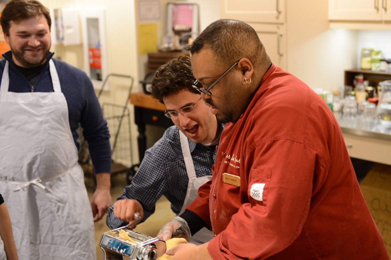 A chef and a participant use a pasta machine while another person watches and smiles in a cooking class.