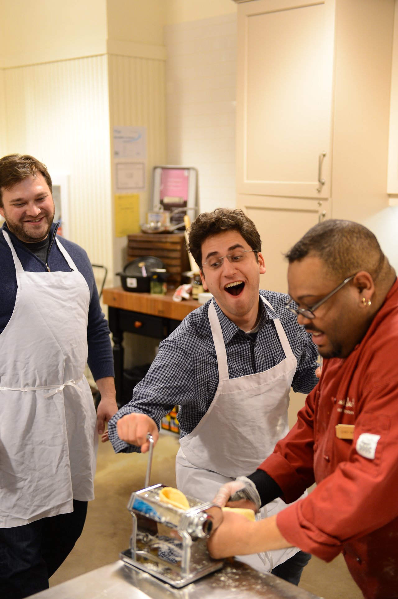 Three people in aprons make pasta using a hand-cranked machine in a cooking class, smiling and engaged.