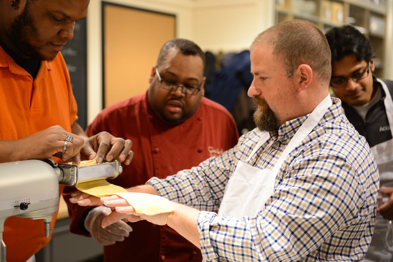 Several people in a cooking class make fresh pasta using a pasta machine, with one person guiding the dough.