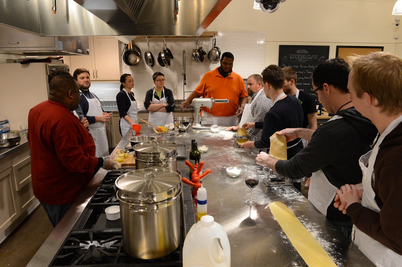 A group of people in aprons participate in a cooking class, rolling out fresh pasta dough on a stainless steel counter.