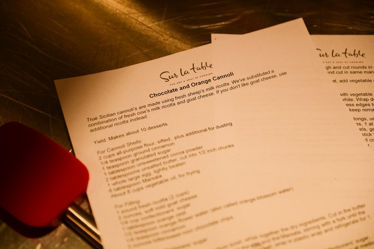 A printed recipe for chocolate and orange cannoli rests on a stainless steel surface with a red spatula nearby.