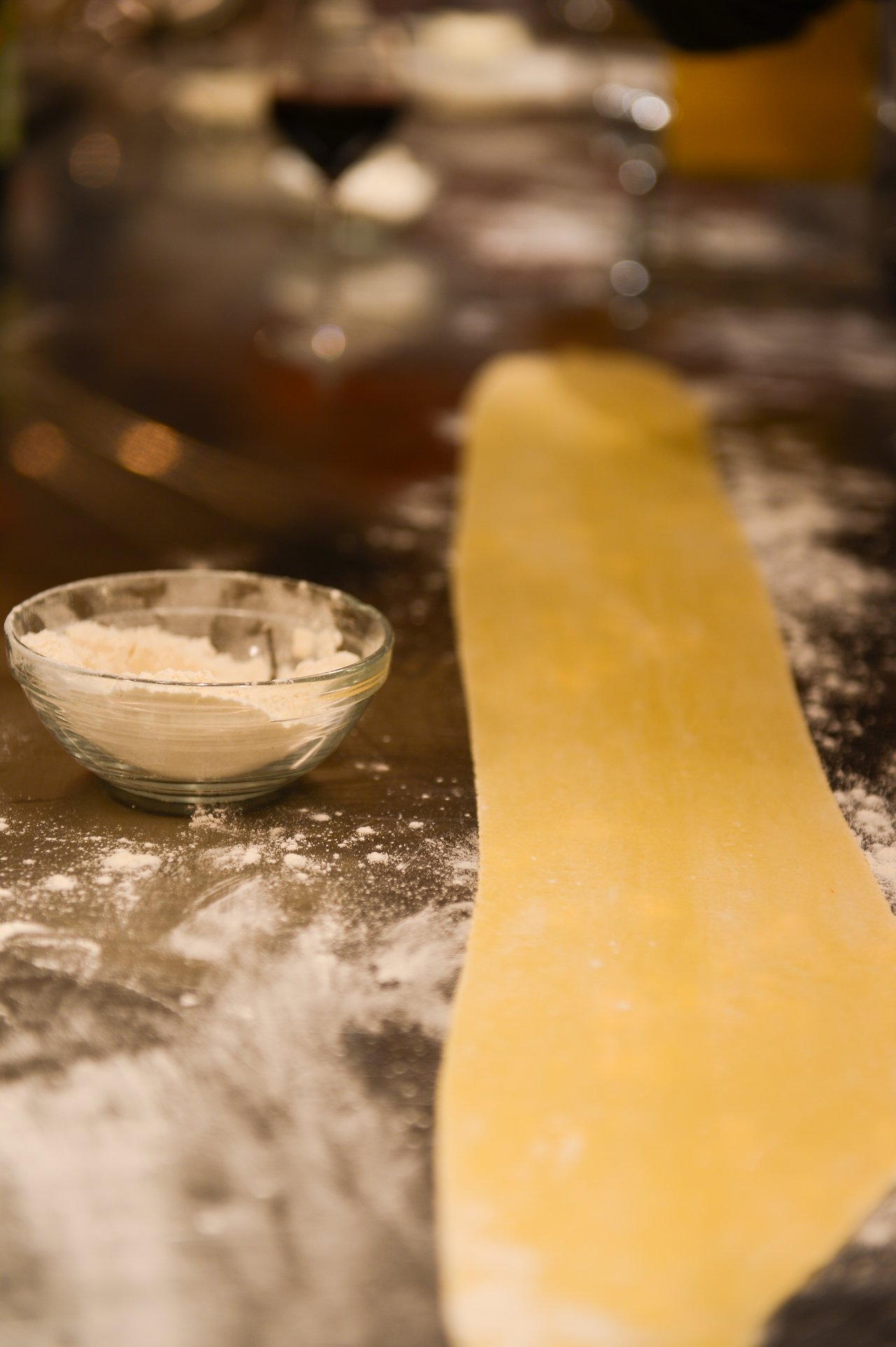 A sheet of rolled-out pasta dough on a floured surface with a small bowl of flour nearby.