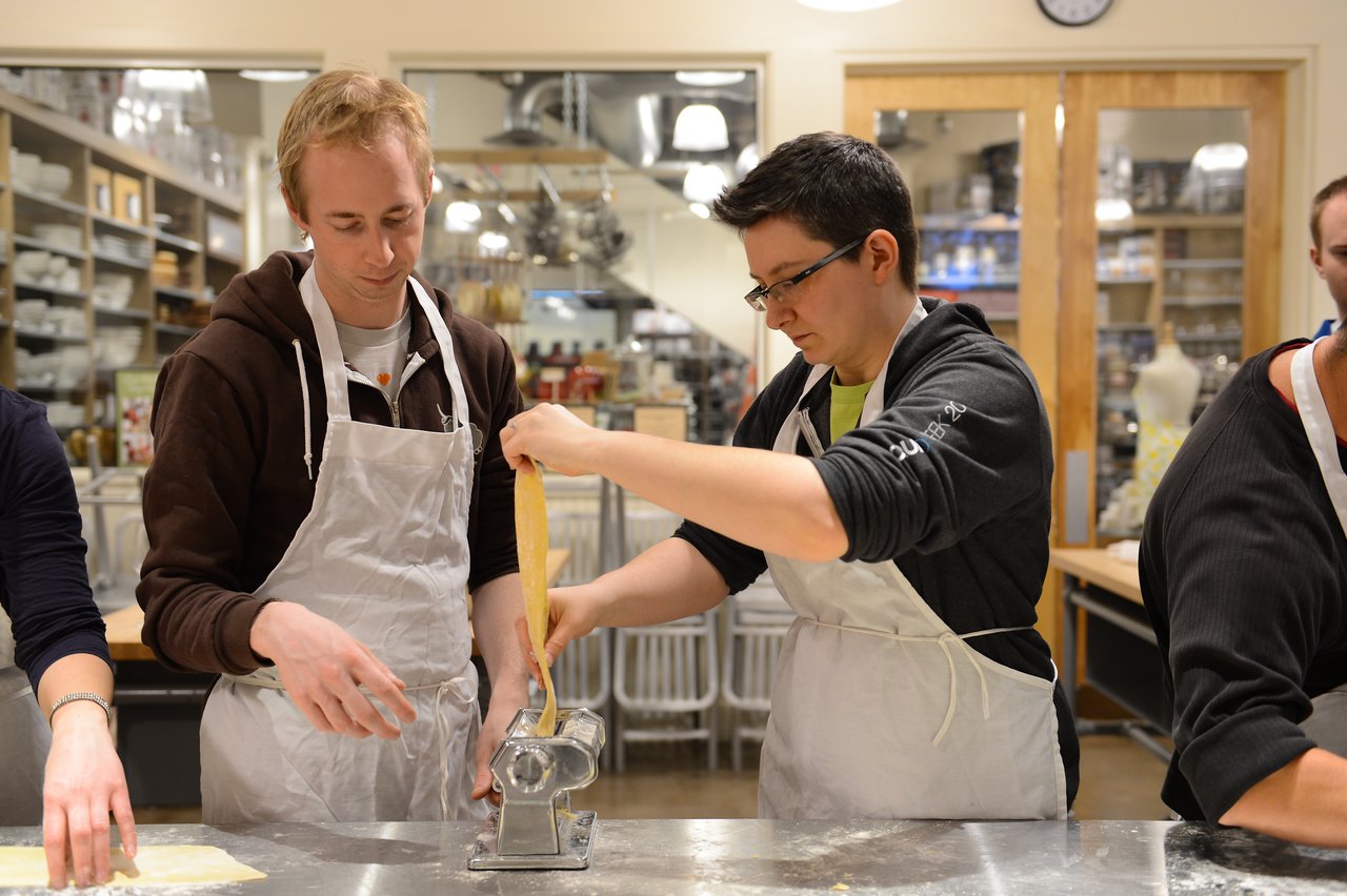 Two people wearing aprons roll out fresh pasta dough using a pasta machine in a cooking class.