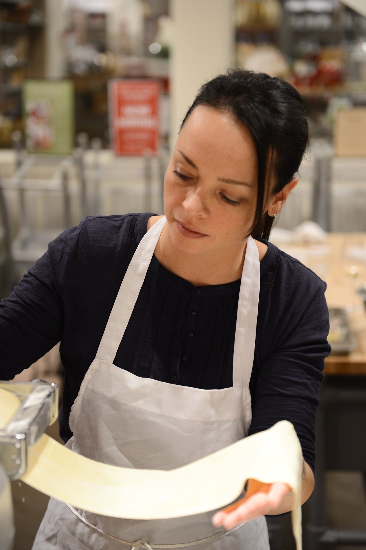 A woman wearing a white apron rolls out fresh pasta dough using a pasta machine in a cooking class.