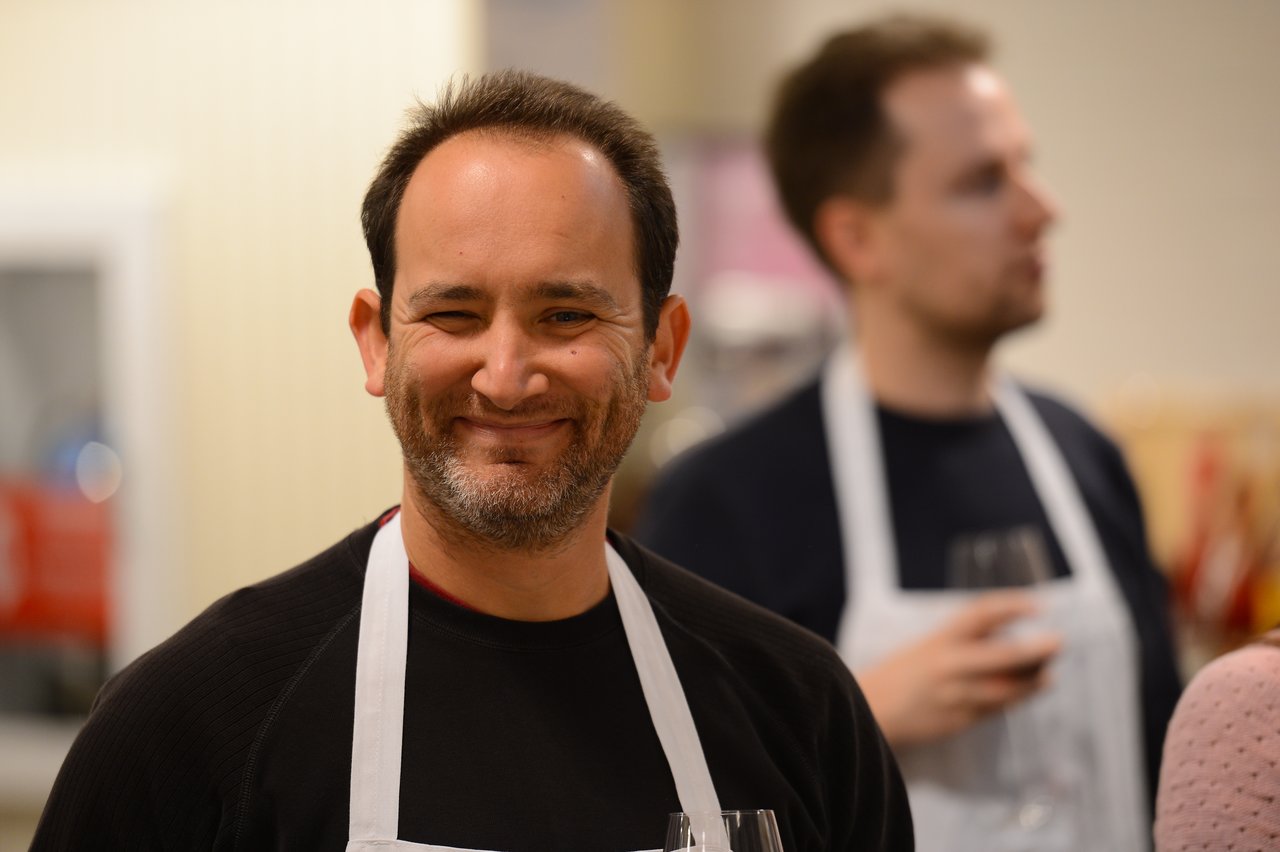 A smiling man in a white apron stands in a cooking class, with another participant holding a glass behind him.