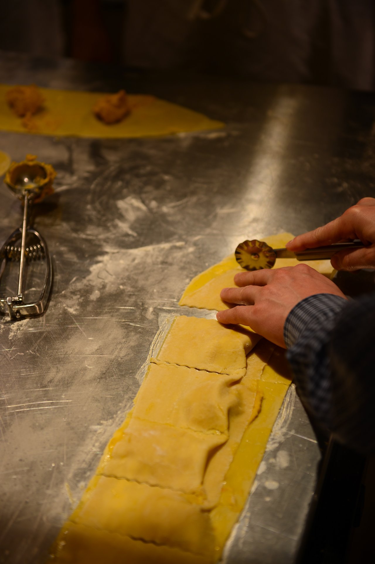 Hands cutting fresh ravioli dough with a fluted pastry cutter on a floured metal surface.