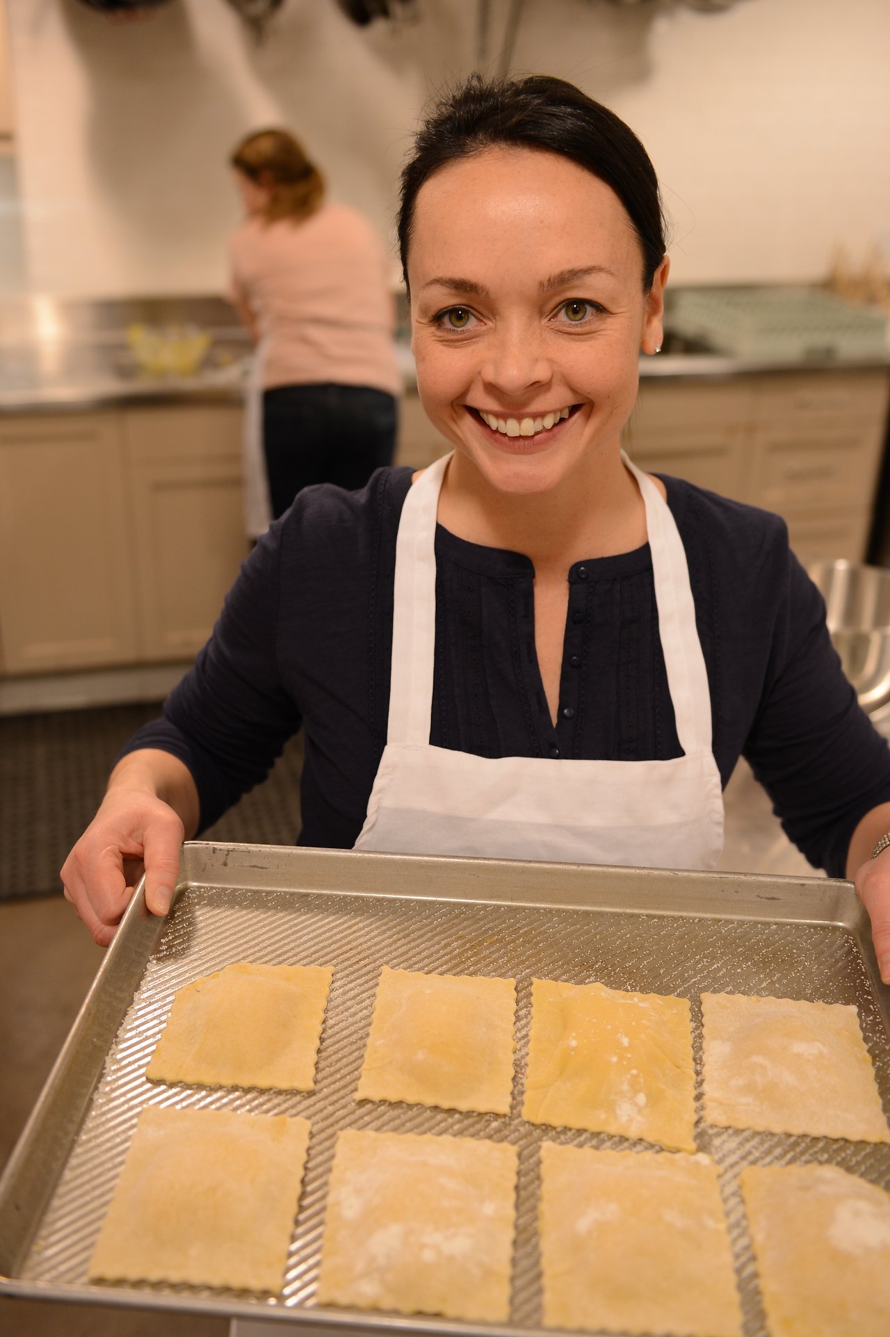 A woman wearing an apron smiles while holding a tray of freshly made ravioli in a kitchen.