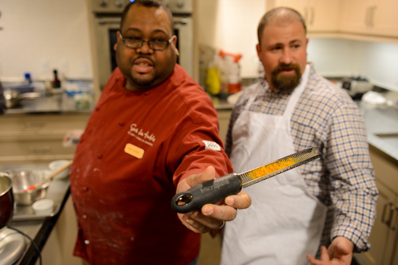 A chef in a red coat holds a zester with grated orange peel while teaching a cooking class.