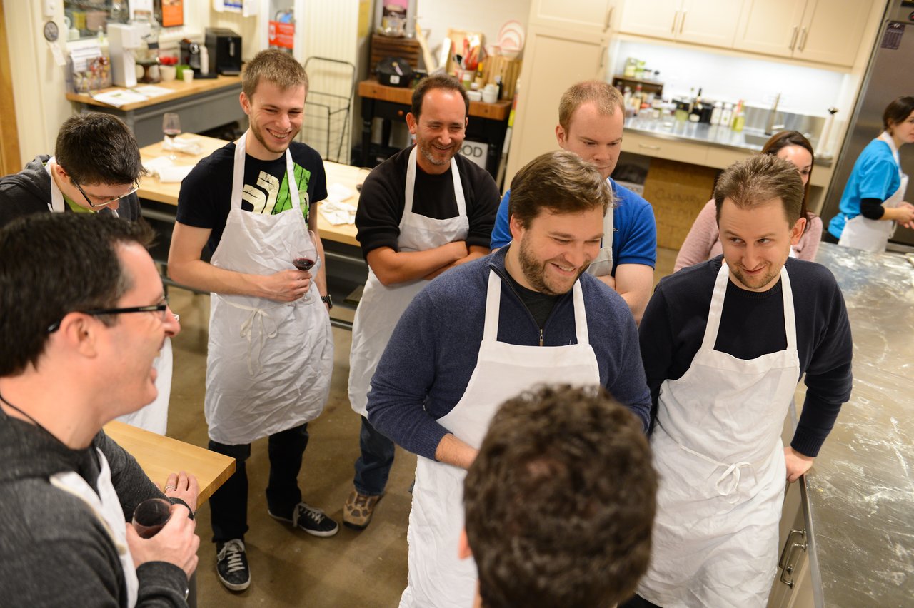 A group of people wearing aprons stand together in a kitchen, smiling and engaged in a cooking class.